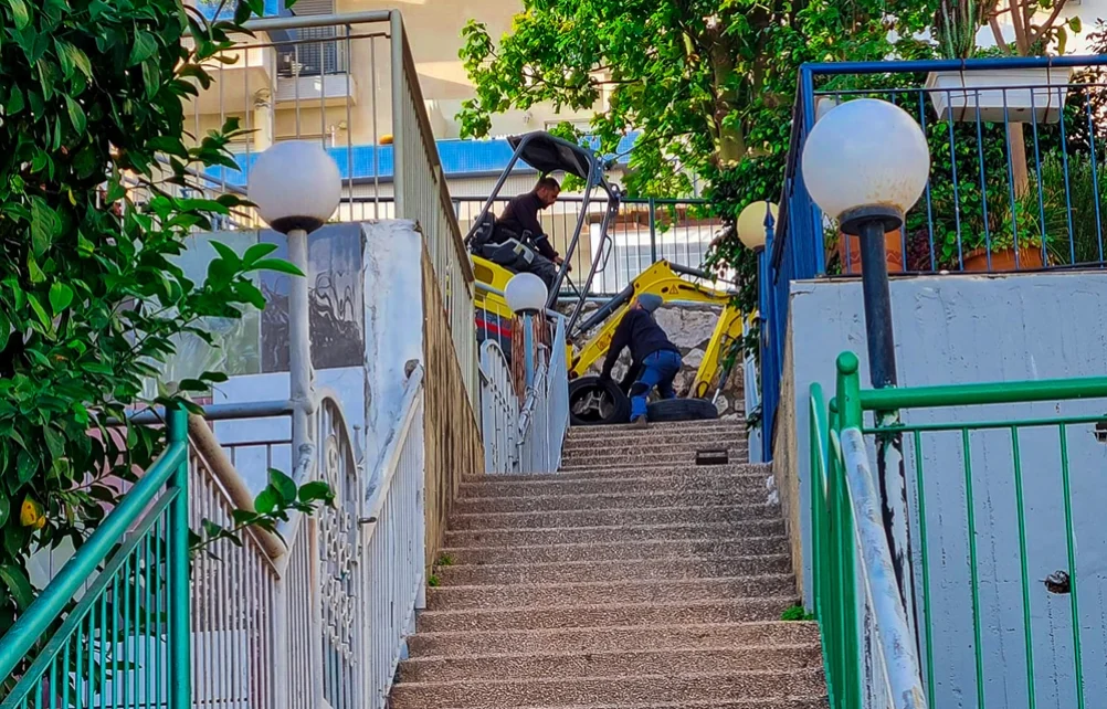 גרם מדרגות חיצוני לצד תאורת רחוב, כשברקע מחפרון קטן עובד במעלה השביל בין המבנים.