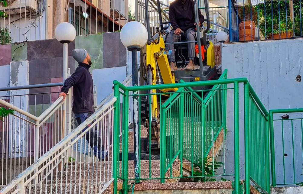 מחפרון קטן מתמרן במעלה גרם המדרגות החיצוני לצד מעקות ירוקים ואביזרי תאורה.